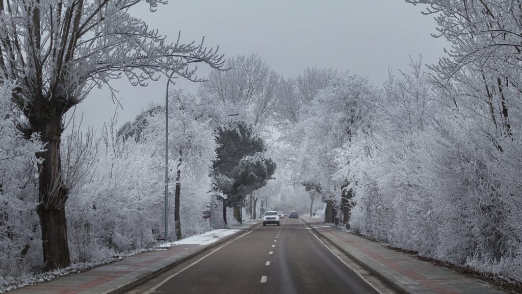 A car drives down a snowy winter road surrounded by frost-coated trees.
